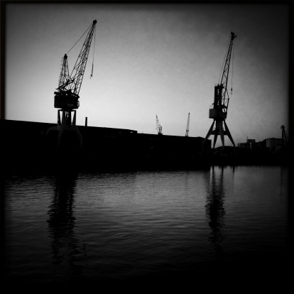 Silhouetted cranes at a harbour in the evening shortly after sunset. The cranes are reflected in the water.