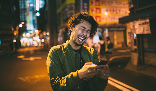 Young japanese man texting on the phone during he walks on the street. There is night and there is nobody on the street. He holds him phone and laughing.