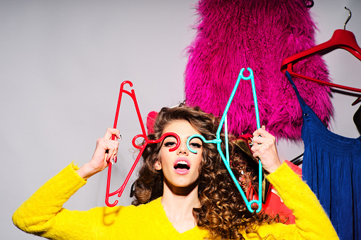 Crazy young girl with curly hair in yellow sweater holding hangers standing amid colorful clothes pink red blue colors on grey wall background, horizontal picture