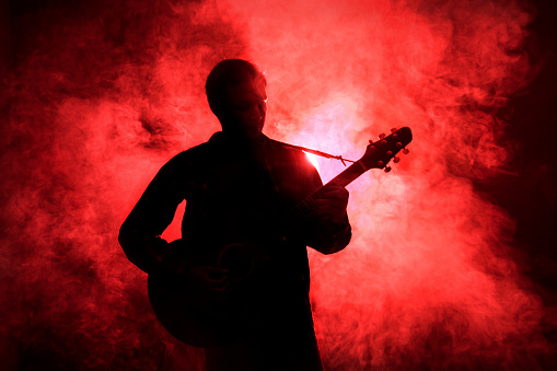Silhouette of an unrecognizable male person playing guitar on stage with red light and smoke