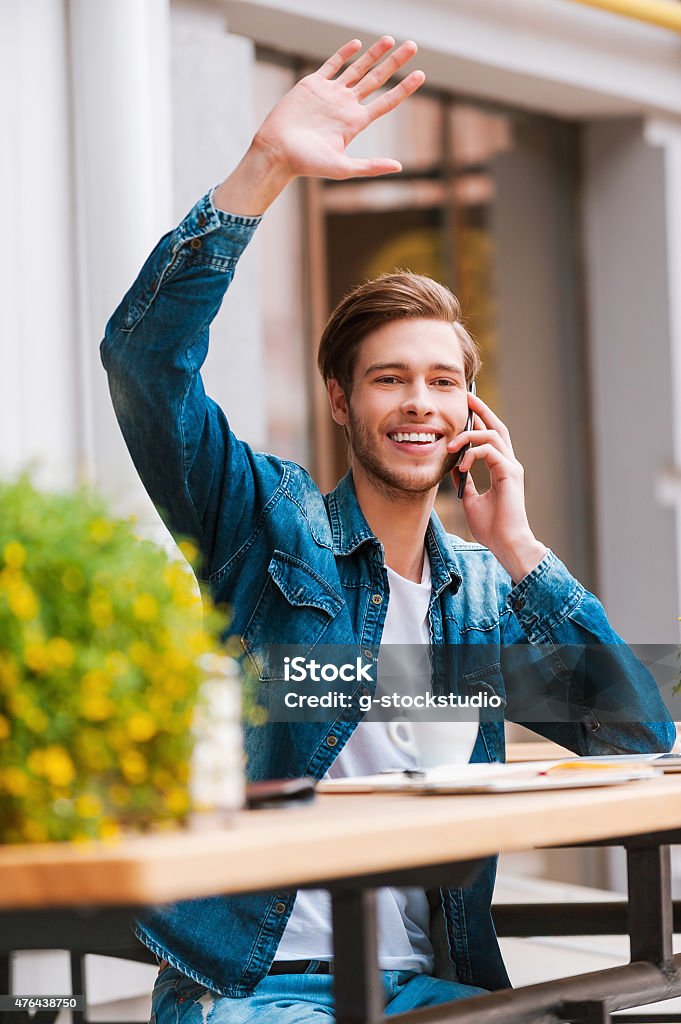 Meeting his friends in cafe. Smiling young man talking on the mobile phone and waving while sitting at sidewalk cafe Men Stock Photo