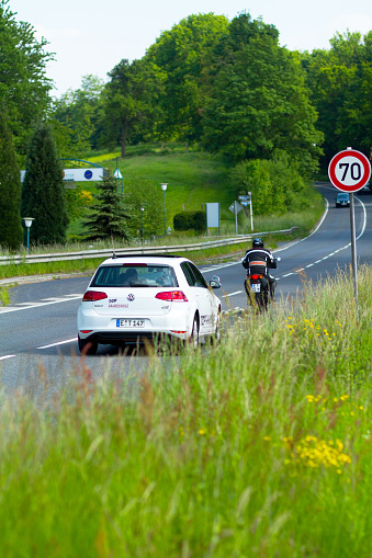 Essen, Germany - May 22, 2015: Driving lesson for motorcycle on road in Ruhrgebiet in spring. Man on motorcycle is driving ahead car of driving school. They are passing road sign for maximum speed 70km. Driving teacher is sitting inside of car.