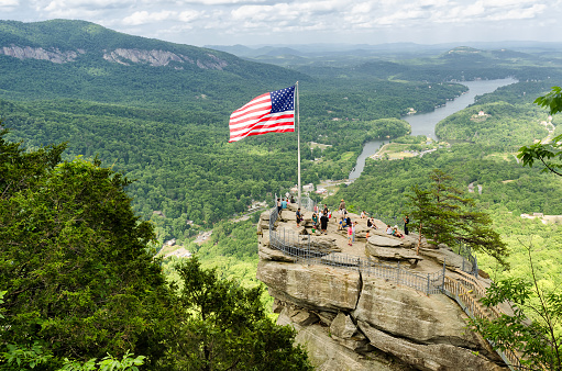 Lake Lure, United States - May 25, 2015: American flag at Chimney Rock mountain, State Park in North Carolina, United States. High point view on chimney rock with people around and Lake lure on a background. 