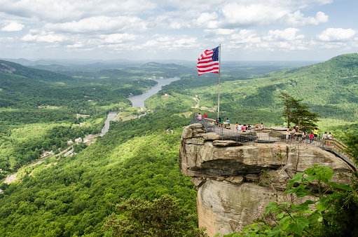 Lake Lure, United States - May 25, 2015: American flag at the top of Chimney Rock mountain, State Park in North Carolina, United States. High point view on chimney rock with people around and Lake lure on a background. 