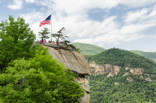 Lake Lure, United States - May 25, 2015: American flag at the top of Chimney Rock mountain, State Park in North Carolina, United States. High point view on chimney rock with people around and mountains on a background. 