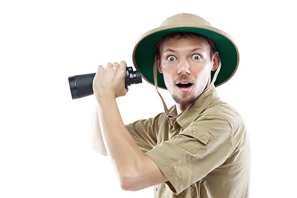 Surprised young man wearing a pith helmet and holding binoculars, isolated on white