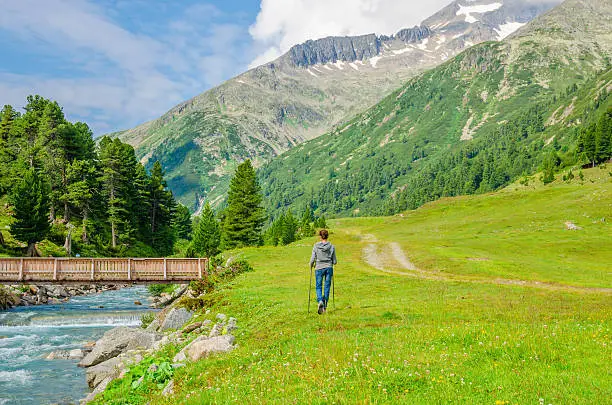 Young woman mountaineer walking over a mountain stream, on a background of green meadows and peaks of the Alps, Austria