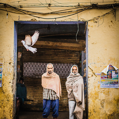 Old Delhi, India - March 1, 2015: On a rainy Spring day, two Indian men standing in a building entrance, upper bodies wrapped in blankets due to cold weather.