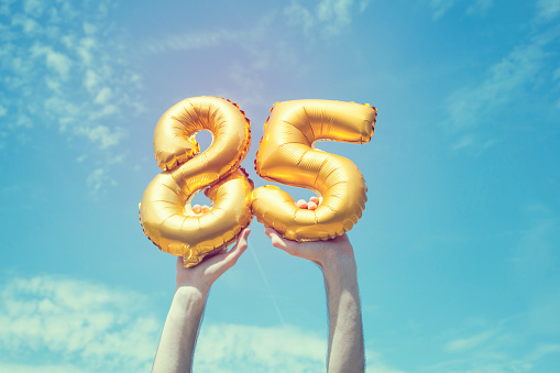 A gold foil number 85 balloon is held high in the air by caucasian male hand.  The image has been taken outdoors on a bright sunny day, the sky is blue with some clouds. A vintage style effects has been added to the image.