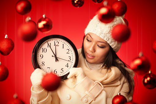 A young Asian Chinese Woman holding up a deadline clock for Christmas holiday shopping. She is holding and staring at the clock for time is running out for the Christmas shopping days. Photographed in red background with Christmas theme decorative setting. 