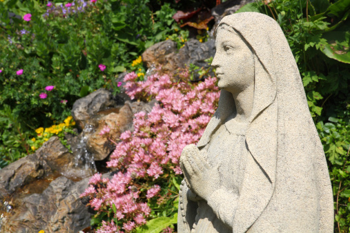 Statue of the Virgin Mary in a garden surrounded by various plants, flowers and a waterfall. With copy space.