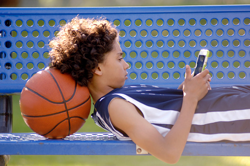 Ten year old African American boy sitting on a blue park bench outdoors, with a basketball, and texting on a mobile tablet.