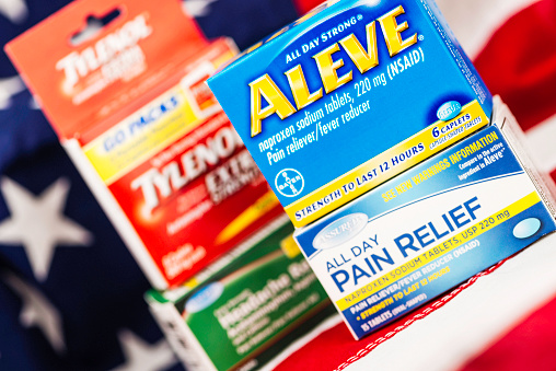 Peyton, Colorado, USA - June 7, 2015: A horizontal studio shot of the name brand pain relief medications, Aleve and Tylenol. Beneath each brand name box, is a generic counterpart with similar ingredients. In the background is an American flag to depict choices in the USA for purchasing generic vs name brand medication.
