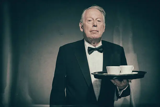Waist Up of Senior Male Butler Wearing Formal Suit and Bow Tie Holding Tray with Two White Coffee Cups in Dimly Lit Studio