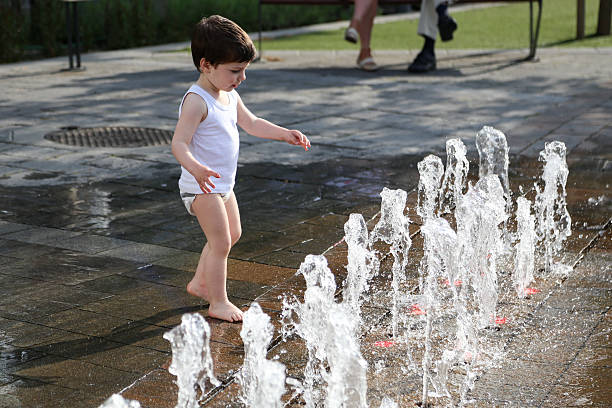 Little blond boy wearing shorts and t-shirt standing barefoot by the water faucet outdoors, washing his toy car, extreme temperatures in the city due to climate change