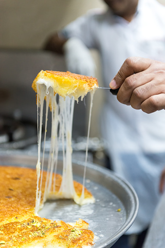 iStockalypse Dubai.  Closeup of a baker’s hand pulling freshly baked Middle Eastern, Palestinian Kunafa pastry made with cow’s milk clarified butter, noodle threads and cheese in a commercial kitchen.  For sale at a bakery in Dubai, UAE, Middle East, GCC.