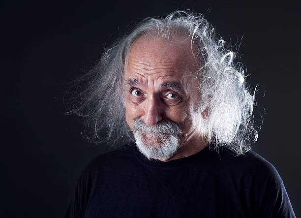 A teacher with a long white beard, big gray hair, and glasses is wearing a checkered shirt and sports jacket in front of a blackboard with complex formulas written on it. He looks sternly at the camera over his glasses with his index finger raised.