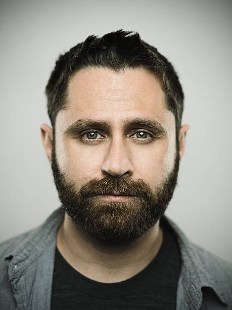 Portrait of a real caucasian young man with neutral expression. Brown eyes and hair with beard. 35 years old north american. Vertical color image from a dslr camera in studio.