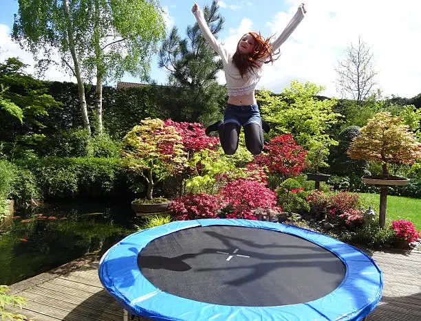 Photo showing a young girl jumping high on her round trampoline, reaching her hands into the air as she bounces.  There is no safety net surrounding the trampoline, so that the garden, pond and decking can be seen more clearly in the photograph, as well as the bouncing girl.