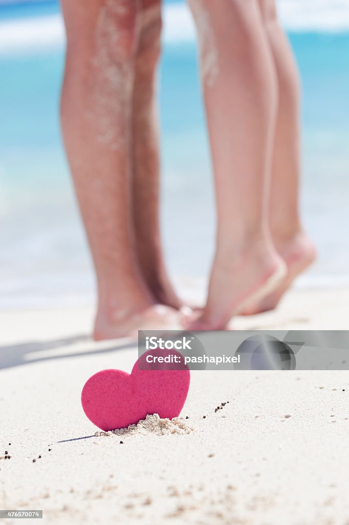 Couple of male and female legs on beach Barefoot female legs standing up tiptoe on man's foots on beach with turquoise sea background, decorated pink heart object.  Romantic honeymoon vacation concept 2015 Stock Photo