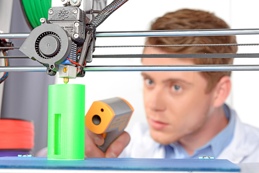 Young handsome scientist watching 3D-printer printing a green plastic model and holding digital thermometer, wearing white lab coat in laboratory