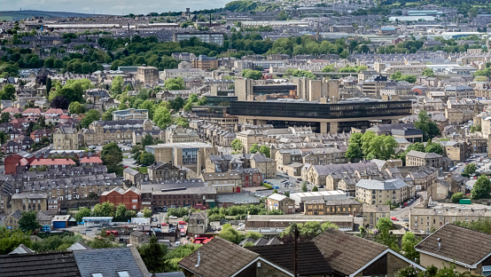 A view of Halifax, West Yorkshire encompassing foreground houses and including the City Center with a famous building society head office in the center of the image. Mills and northern type buildings can also be seen. Green trees are also in the image as are the distant moors, green belt area and wind turbines in the extreme background.