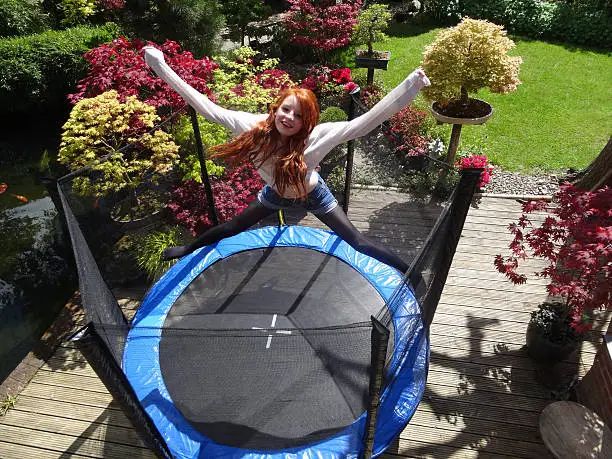 Photo showing a girl jumping extremely high on the trampoline, bouncing so high that she is almost above the surrounding safety net.  The trampoline is pictured positioned on wooden decking, which forms the main patio area in this landscaped garden, planted with maples (acers) and flowering azaleas.