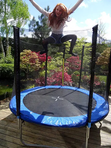 Photo showing a girl playing outside in her back garden, doing some high jumps and bouncing on her new round trampoline, which comes complete with the obligatory safety net and is positioned on timber decking.  The colourful garden behind is a mixture of Japanese maples and azaleas, along with a koi pond.