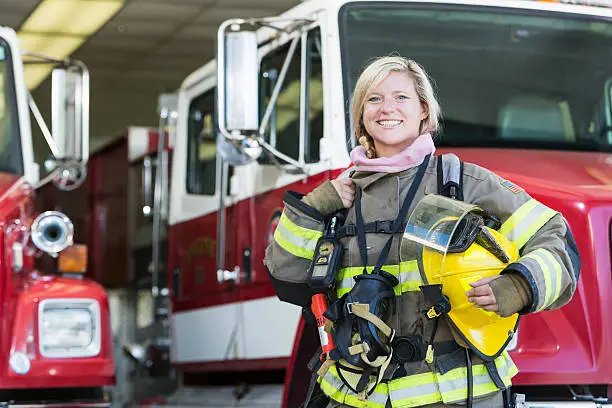 A young, smiling, female firefighter standing in front of fire trucks at the station, wearing a protective suit, ready to respond to an emergency. She has an American flag patch on her sleeve.