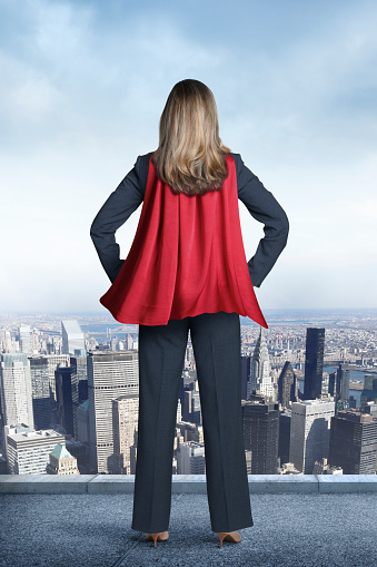 A rear view of a businesswoman wearing a red cape  looking out over the city below her. She is standing her hands on her hips as a gentle breeze flows through the cape  A blue sky and patchy clouds are in the distance. 