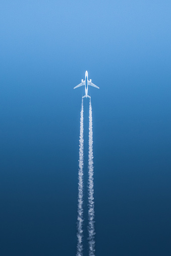 A white jet airplane with blue center, as seen from underneath, in flight with white contrails against a deep blue contrast background.