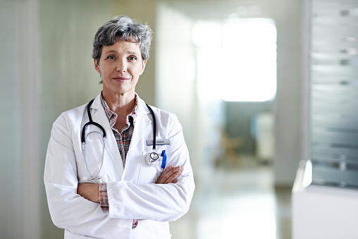 Portrait of healthcare worker wearing protective workwear during coronavirus epidemic. Mature female surgeon is standing with arms crossed. She is against white background.