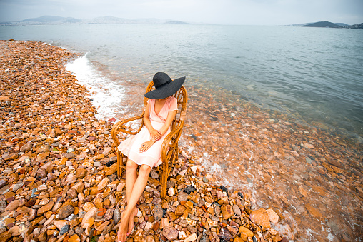 Young woman in pink dress and big black hat resting in the wicker chair on the beach in cloudy weather. 