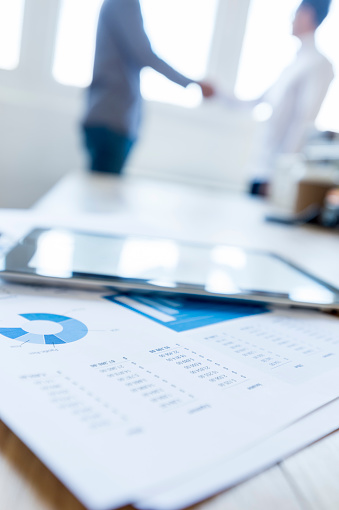 Two businessmen are shaking hands in the background. There are charts, graphs and a digital tablet in the foreground. Focus is on the foreground with back lit people in the background. They look like they have just done a business deal. Financial figures are also visible.