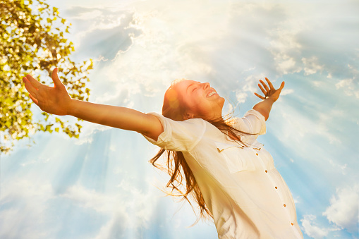 Pretty young woman raising her arms and enjoying perfect sunny day in nature. Her hair is long and bouncy, wearing a white shirt and enjoys smiling with eyes closed. Above her it is the sky with clouds through which spread sun rays. AdobeRGB color space. Small amount of grain added intentionally for better final impression.