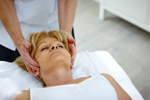 Mature woman lying on table receiving head massage from osteopath therapist