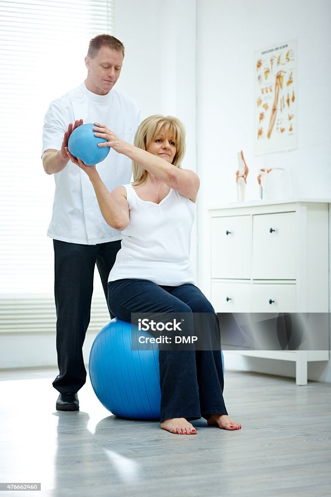 Physical therapist assisting senior woman at medical gym Senior woman sitting on fitness ball holding fitness ball being assisted by her physiotherapist in medical room Physical Therapy Stock Photo