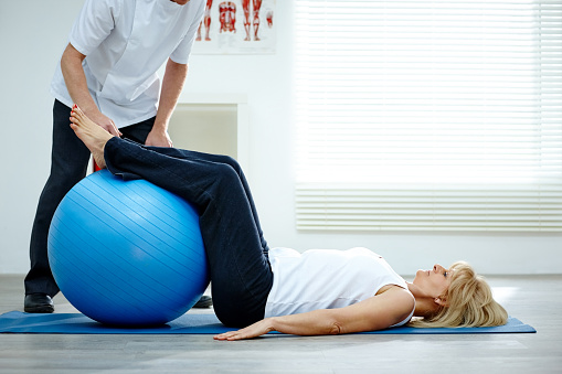 Physical therapist working with a senior woman lying on exercise mat with her legs on pilates ball in gym at hospital