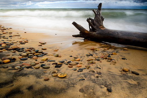 Long shutter speed was used to blur the waves crashing into the shore of Lake Superior at Whitefish Point in Paradise Michigan.  A large log of drift wood is being battered by the waves and the water is flowing back into the lake over the sand and multi colored rocks.  Shot with a tilt shift lens and a high resolution D800.