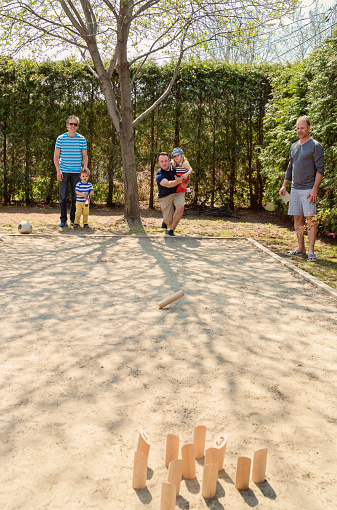 Three dads and two little kids are playing Molkky, a wood skittles game in a suburb backyard. This game was create in Finland in 1996, and is gaining popularity in all parts of the world, like here in a small North American city. Vertical full length shot on a very sunny day.