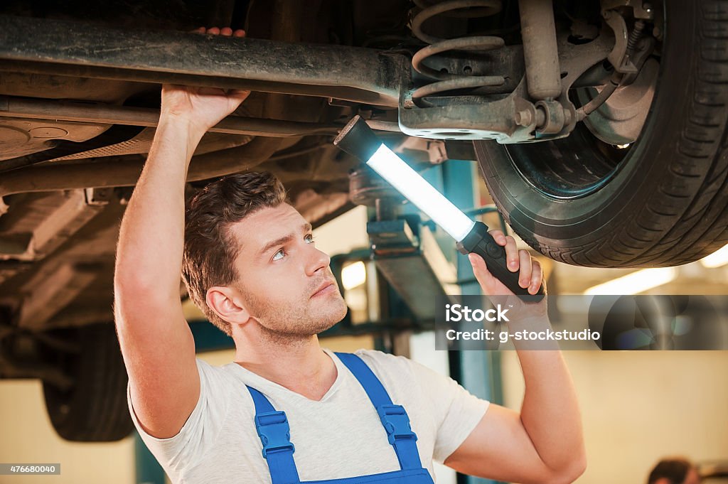 Checking every detail for defects. Confident young man in uniform holding lamp while standing underneath a car in workshop 2015 Stock Photo