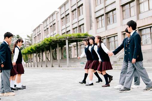A view of a Japanese high school. A group of students exercise together in the playground, outside their school building, Exterior, smiling, playing, hand in hand, contemporary architecture, exterior view, horizontal composition. 