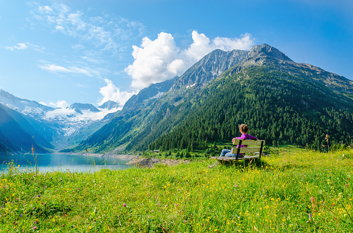 A young woman sits on bench beside an azure mountain lake on the background of the high peaks of the Alps, Zillertal, Austria