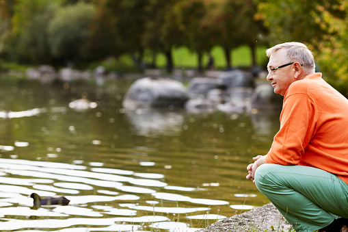 Profile of aged man in glasses sitting near pond in park watching ducks