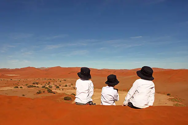 Three little boys wearing white shirts and identical dark blue sun hats are sitting on top of red sand dunes at Sossusvlei in Namibia. The children are here seen from the back as they are looking at a beautiful view over the Sossusvlei valley and the surrounding dunes from the top of the 'Big Mama' dune.