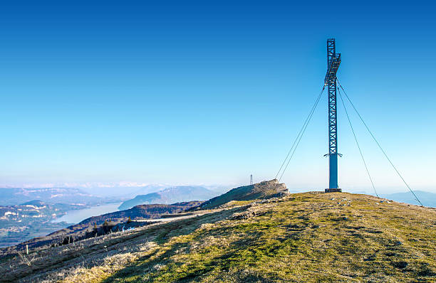 Scenic horizontal photography of the Grand Colombier mountain top, rolling landscape, with a metal cross at the top of the hill. Grass meadow hill mountain top, scenics landscape view from top mountain peak of Alps chain in summer season. Photography taken on famous local landmark Grand Colombier mountain (near Culoz city, and close to the border with the Haute-Savoie), in Bugey, in Ain, Rhone-alpes region in France (Europe). Color image with a big metallic cross shape at the top of the hill.