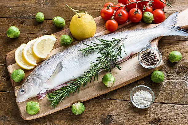 Fresh fish bass garnished rosemary on wooden board. Brussels sprouts and cherry tomatoes for garnish. Spices, like lemon, salt and pepper.