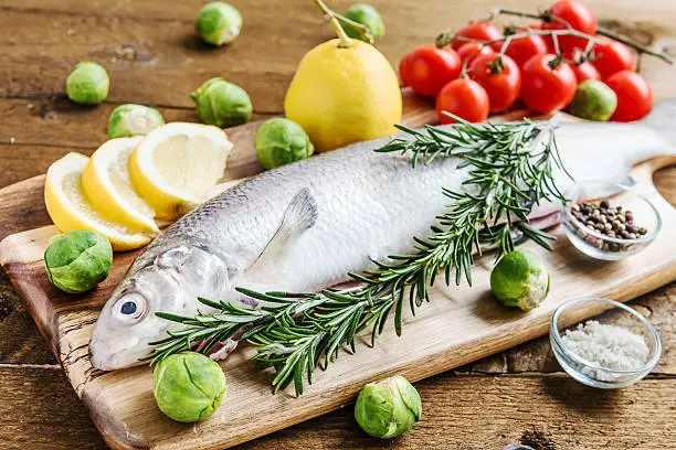 Fresh fish bass garnished rosemary on wooden board. Brussels sprouts and cherry tomatoes for garnish. Spices, like lemon, salt and pepper.