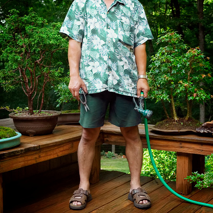 A man standing next to his bonsai  trees holding garden tools.