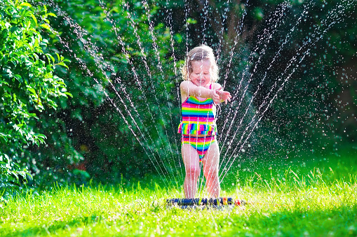 Child playing with garden sprinkler. Kid in bathing suit running and jumping. Kids gardening. Summer outdoor water fun. Children play with gardening hose watering flowers.